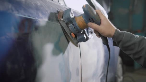 Trabajador está cepillando el tinte azul viejo del cuerpo del automóvil usando amoladora manual, reparando para pintar en un taller de coches — Vídeo de stock