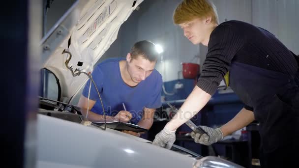 Maestro está inspeccionando los sistemas internos del automóvil en un taller de carrocería del coche, asistente está escribiendo resultados en una hoja de papel — Vídeos de Stock
