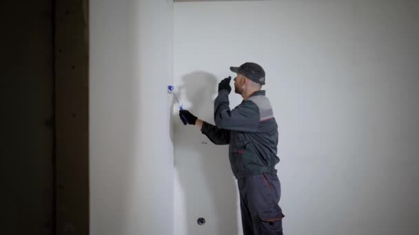 Ein junger Mann in Bauuniform bemalt die Wände in der Wohnung, der Bauarbeiter arbeitet als Walze — Stockvideo