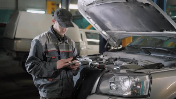 El reparador toma notas en la tableta, el hombre sostiene un gadget en su mano y va a arreglar el coche — Vídeos de Stock