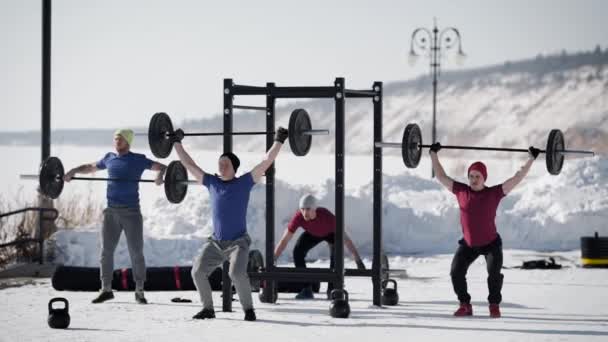 Cuatro levantadores de pesas se entrena en un día de invierno al aire libre, levantando pesadas varillas, de pie en una zona deportiva nevada — Vídeos de Stock
