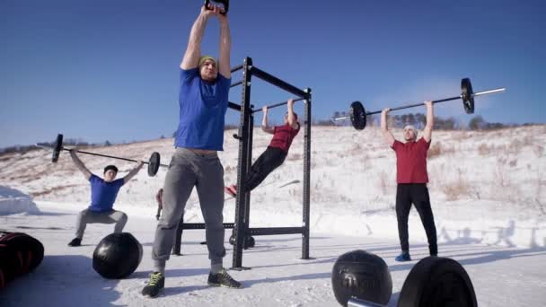 Eine Gruppe von vier jungen Sportlern trainiert an einem Wintertag auf einem Sportgelände, einer zieht am Balken, der zweite hebt die Kettlebell, zwei heben die Stangen. — Stockvideo