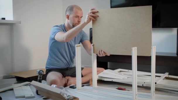 Вмілий чоловік встановлює деталі нового столу в кімнаті, що сидить на підлозі між частинами, картоном та інструментами — стокове відео