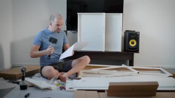 Homem está olhando para uma instruções para a montagem de móveis, segurando martelo, em seguida, girando livro e sorrindo para a câmera, sentado em um piso — Vídeo de Stock