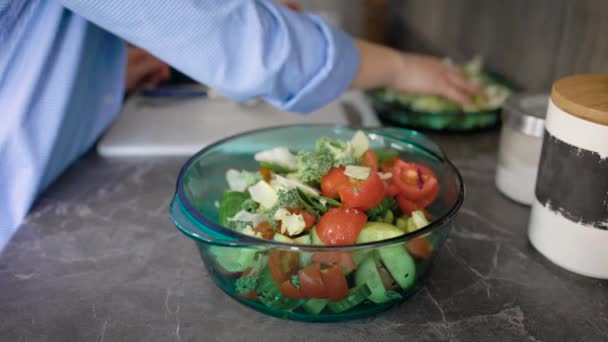 As mãos femininas estão colocando folhas de alface cortadas em uma tigela de salada com tomates e pepinos em uma mesa de cozinha — Vídeo de Stock