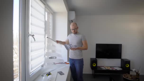 Homem está lendo uma instrução de instalação para cortinas nas janelas e olhando para detalhes, de pé na sala de estar de sua casa — Vídeo de Stock