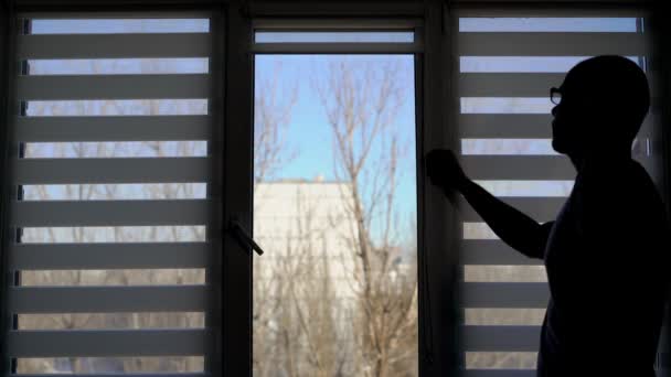 Человек приближается к окнам внутри дома против света и падение белых жалюзи, осенний пейзаж день на заднем плане — стоковое видео