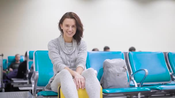 陽気なブルネットの少女は空港で彼女の飛行を待っている袋の上に座って彼女の将来の休暇と旅行を楽しんで — ストック動画