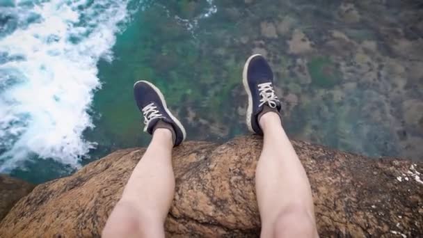 Benen van de mens opgestart in donker blauwe sneakers zijn opknoping van de rand van de klif over krachtige golven hieronder — Stockvideo
