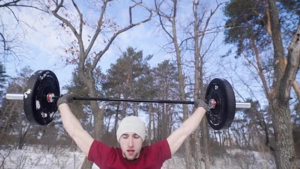 A young man who is engaged in winter time on nature crouches with a heavy barbell, the person looks sporty and healthy — Stock Video