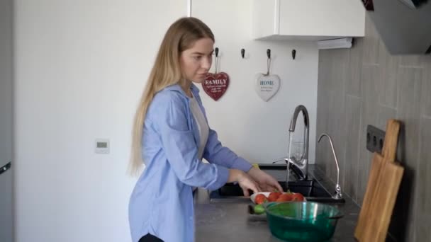 Uma jovem mulher lava legumes frescos para fazer uma salada leve de tomates e pepinos, que ela vai cortar — Vídeo de Stock