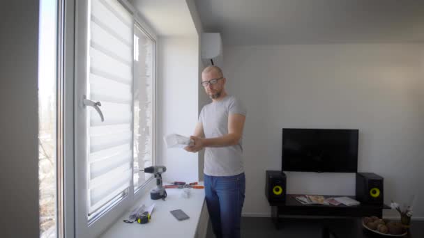 El hombre adulto desempaca el embalaje de las cortinas, cuelga a la gente en la ventana con protección solar en la sala de estar — Vídeo de stock