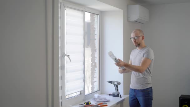 Взрослый мужчина делает ремонт в своей квартире, человек распаковывает занавески, которые он вешает на окно — стоковое видео