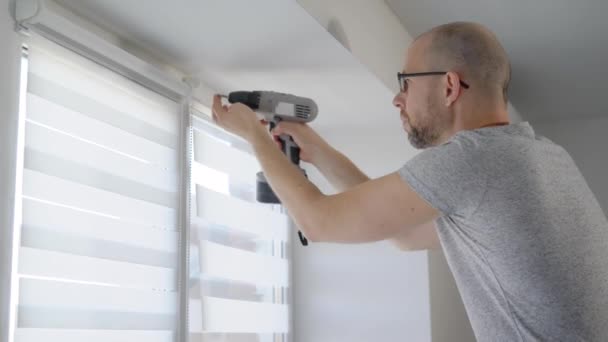 Een volwassen man een elektrische boor in zijn handen houdt en schroeven het venster dat hem beschermt tegen licht — Stockvideo