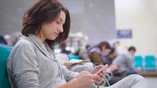 Una mujer bonita se apoya en su mano y escanea las redes sociales en un teléfono inteligente, ella está en el aeropuerto — Vídeo de stock