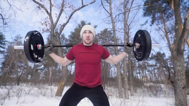 Young handsome bodybuilder doing knee bending exercise with barbell in snowy woods — Stock Video