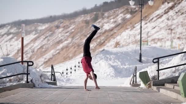 Vista lateral do jovem homem acrobático muscular em pé sobre as mãos e equilibrando na estrada no inverno — Vídeo de Stock