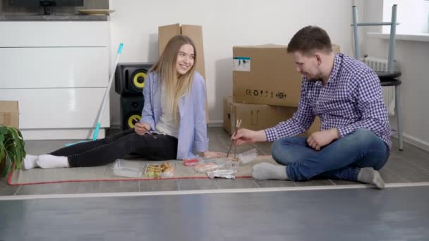 Encantadora esposa y marido guapo están sentados en el suelo en un apartamento vacío y comer comida rápida juntos . — Vídeos de Stock