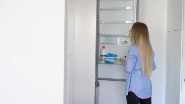 Vista lateral da jovem mulher bonita de pé na cozinha enquanto olha e levando produtos da geladeira — Vídeo de Stock