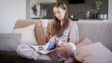 güzel esmer kız ipek giyen pijama oturuyor bir kanepe oturma odasında akşam, çay içme ve dergi okuma