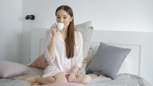 Sozinha menina romântica está sentada em sua cama de manhã, bebendo café de pequeno copo branco e sonhando — Vídeo de Stock