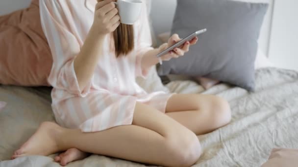 Waking up brunette girl is drinking coffee in her bed in morning time in holidays and typing on her smartphone — Stock Video