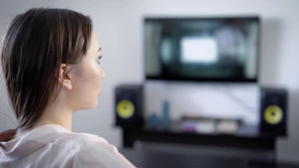 Alone girl is watching tv sitting in a living room of her house in evening, concentrated following the plot of movie, view through shoulder — Stock Video