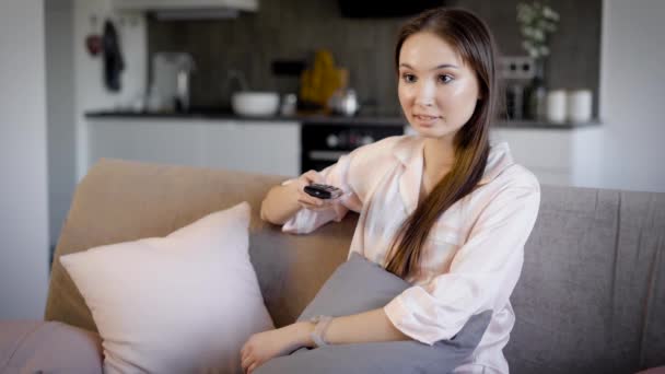 Mulher morena bonita está descansando em um sofá na sala de estar em férias e alternando canais de tv por controle remoto — Vídeo de Stock