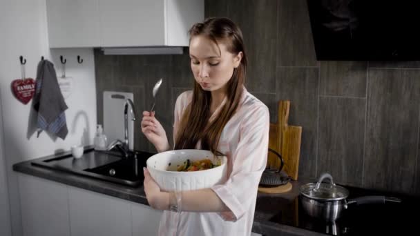 Joven mujer está comiendo ensalada de verduras por cuchara, sosteniendo gran ensaladera blanca en las manos, de pie en una cocina por la noche — Vídeos de Stock