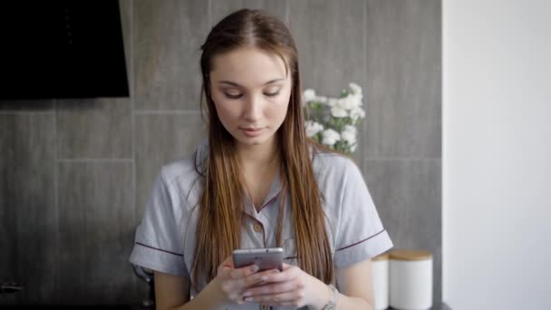 Mujer joven tranquila está escribiendo texto en una pantalla táctil de su teléfono móvil, de pie en la habitación de la casa durante el día — Vídeo de stock