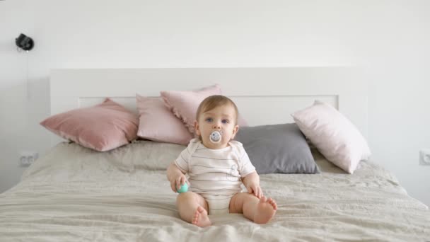 Blondynka blue eyed niemowlę siedzi w duże łóżko z noworodek pacyfikator i oczekuje się w ciągu dnia, w przytulnym domu — Wideo stockowe