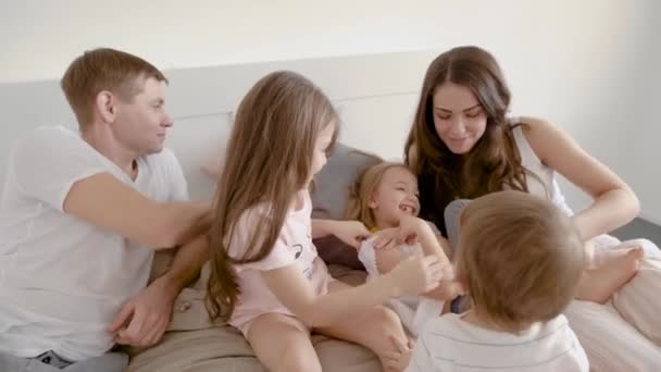 Drie kinderen meisjes spelen en kietelen elkaar in de slaapkamer van de ouders, moeder en vader zijn zittend op bed in de buurt van hen — Stockvideo