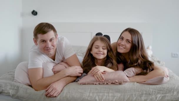 Happy little girl is lying on pillow on bed between her father and mother, mom is kissing daughter in cheek in home — Stock Video