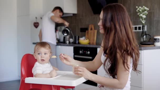 Bruna si nutre di purea di frutta il suo bambino seduto su una sedia in cucina al mattino, suo marito sta cucinando il caffè — Video Stock