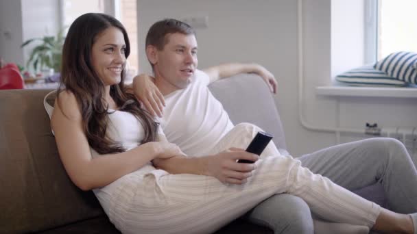 Wife and husband are talking cheerfully sitting on a couch in their living room in nonworking day, man is holding remote control of tv — Stock Video