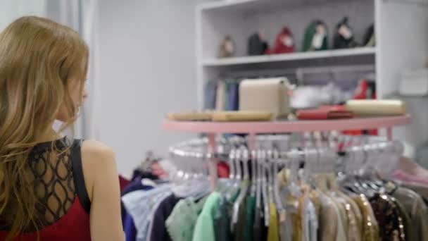Felnőtt nő, piros hullámos haj megy át a csarnok a ruházati bolt, gondolkodás, és keresi a legjobb ruha — Stock videók