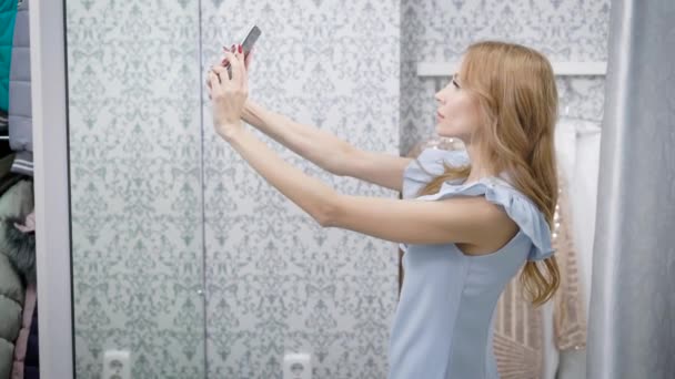 Mujer joven está tomando una foto de su reflejo en un espejo de probador de la tienda de ropa, utilizando el teléfono inteligente — Vídeos de Stock