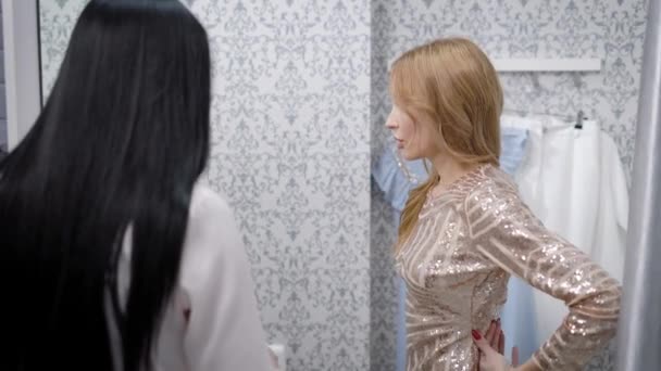 blond dívka se snaží lesklé šaty v šatna, při pohledu do zrcadla a radí se svým přítelem, bruneta