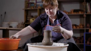 Orta yaşlı kadın seramik vazo karanlık el sanatları atölye çalışmasında, oturan potters tekerleğini kullanarak eller ıslatma yapıyor