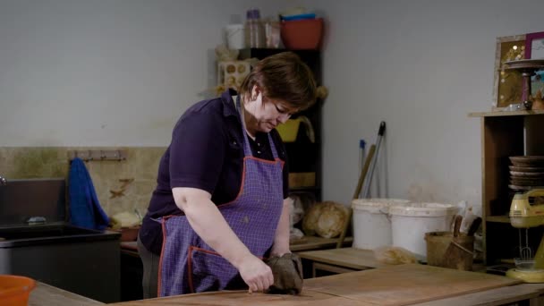 Mujer adulta está usando un delantal está cortando pieza de arcilla por hilo en una mesa de trabajo en un estudio de cerámica — Vídeo de stock