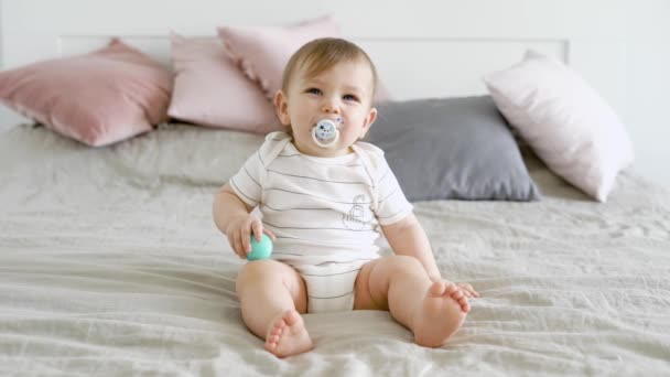 Adorable child with beautiful blue eyes is sitting on the bed sucking on the dummy. — Stock Video