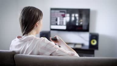 genç kadın oturma odasında kanepede oturup Tv izlerken uzaktan kumanda ile kanal geçiş yapar