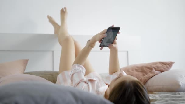 Een jonge vrouw, chatten met vrienden in sociale netwerken houden van een mobiele telefoon in haar handen, ligt ze in bed in de ochtend — Stockvideo