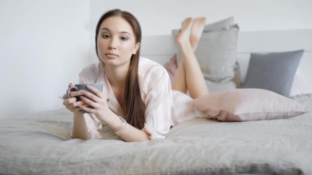 A portrait of a young and pretty lady who drinks a hot drink in the bed, the lady holds a cup and is dressed in pajamas — Stock Video