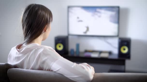 Mujer joven viendo la televisión sentada en el sofá en la sala de estar, ella es aficionada al cine, género de thriller de películas — Vídeos de Stock