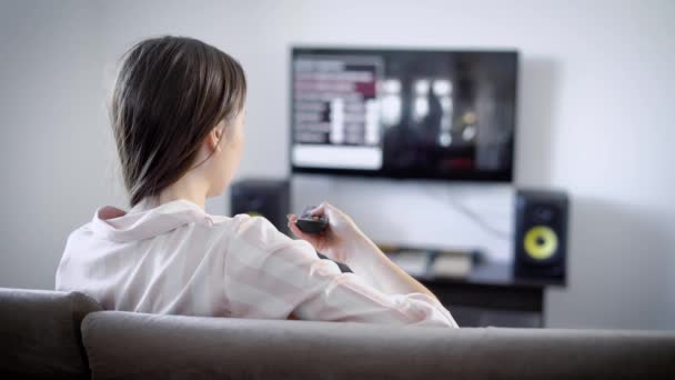 Молодая женщина смотрит телевизор, сидя на диване в гостиной, она переключает каналы с помощью пульта дистанционного управления — стоковое видео