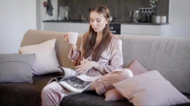genç kadın kanepede oturan ve moda dergisi izliyor ve sıcak çay içme ev yapımı pijama giymiş