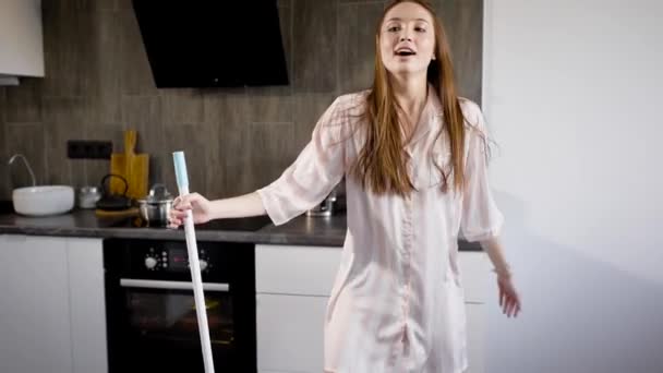 Uma jovem vestida de pijama tolos durante uma dança com uma esfregona na cozinha, a senhora canta canções — Vídeo de Stock