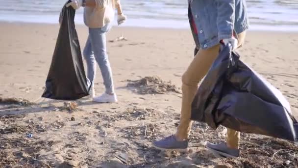 Dos voluntarias están caminando en la playa de arena en el día de primavera y recogiendo los residuos del suelo, poner en bolsas de plástico — Vídeos de Stock