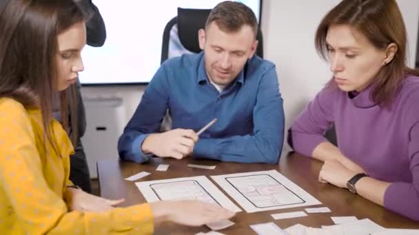 Мужчина и две женщины разрабатывают новое приложение для смартфона, заполняя схемы на бумаге в офисе, сидя за столом — стоковое видео